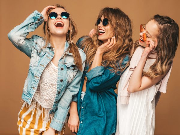 Three young beautiful smiling hipster girls in trendy summer casual jeans clothes. Sexy carefree women posing near golden wall. Positive models going crazy in sunglasses