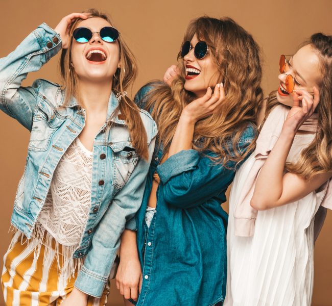 Three young beautiful smiling hipster girls in trendy summer casual jeans clothes. Sexy carefree women posing near golden wall. Positive models going crazy in sunglasses